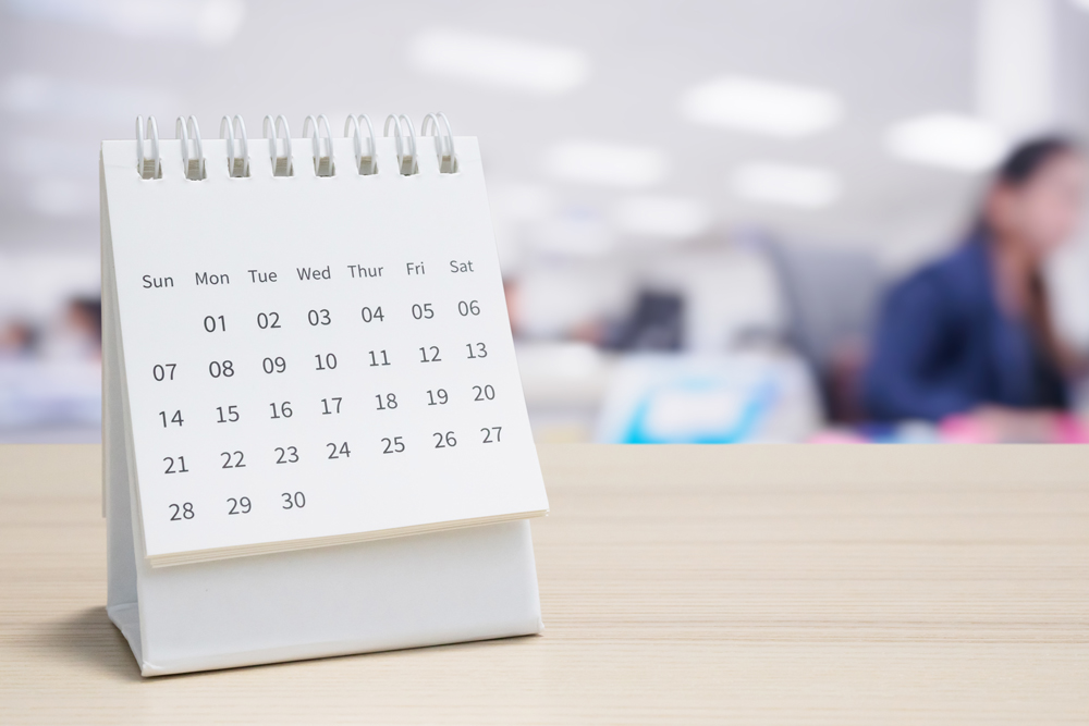 White paper desk calendar on wood table top with blurred office interior background