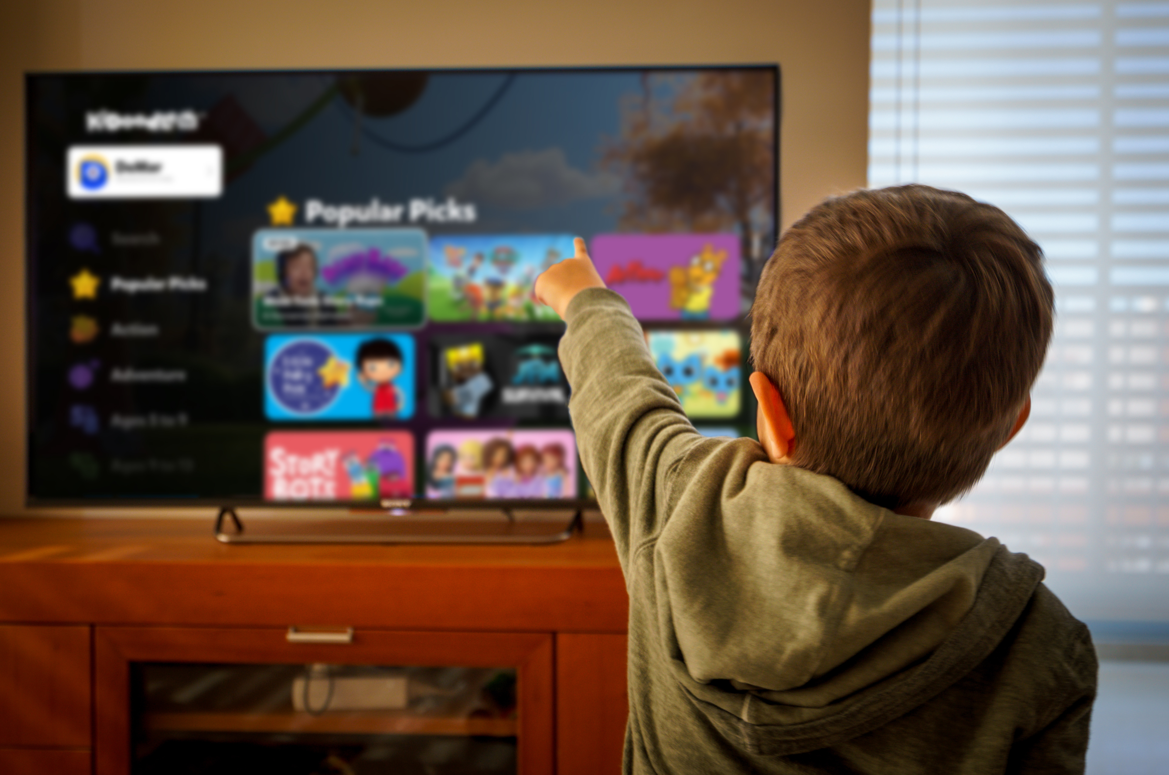 A child in a green hoodie points to a TV displaying the Kidoodle user interface