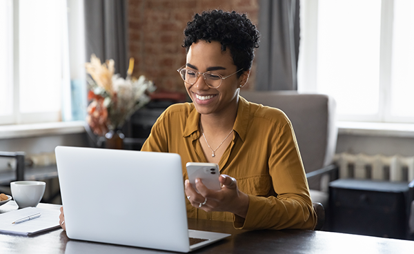 African woman sit at workplace desk holds cellphone staring at laptop, synchronize data between computer and gadget in office, use corporate devices and business application, plan work, use organizer; Shutterstock ID 2159105479; purchase_order: Delivery Hero Innovator page; job: AWS; client: gcrp