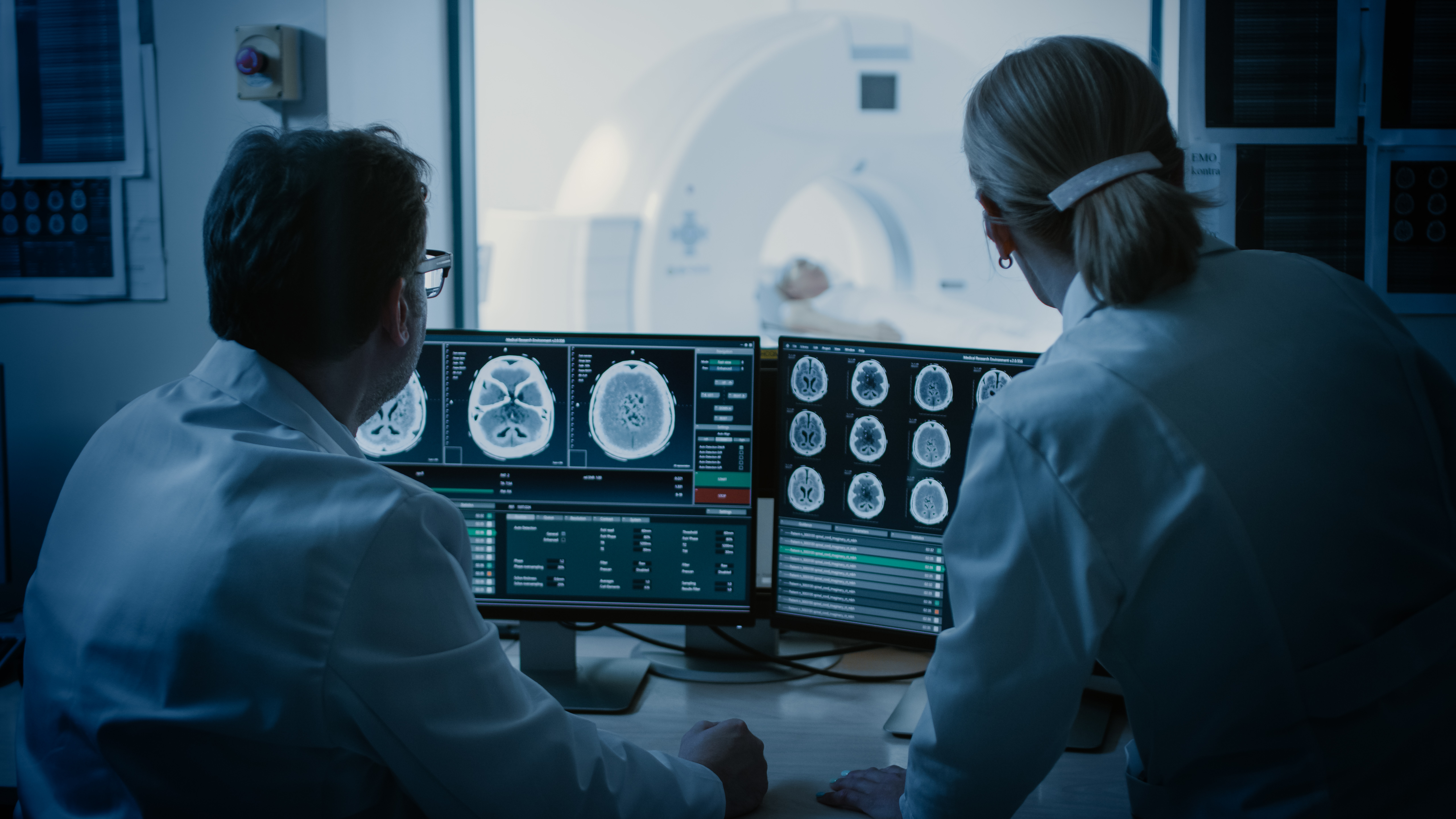 In Control Room Doctor and Radiologist Discuss Diagnosis while Watching Procedure and Monitors Showing Brain Scans Results, In the Background Patient Undergoes MRI or CT Scan Procedure.