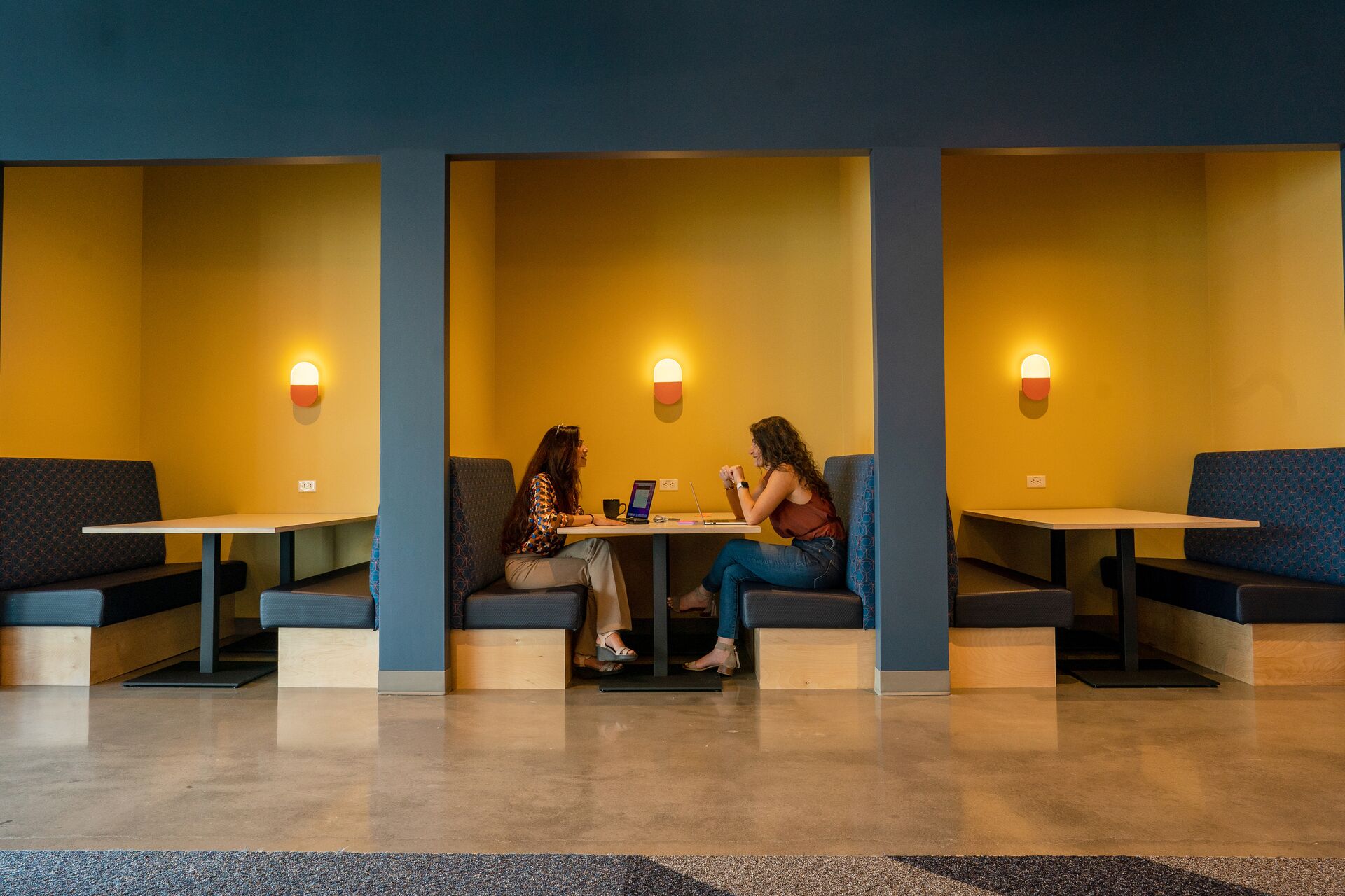 Two women sit in a booth in an Amazon office building with their laptops, appearing to discuss work-related topics.