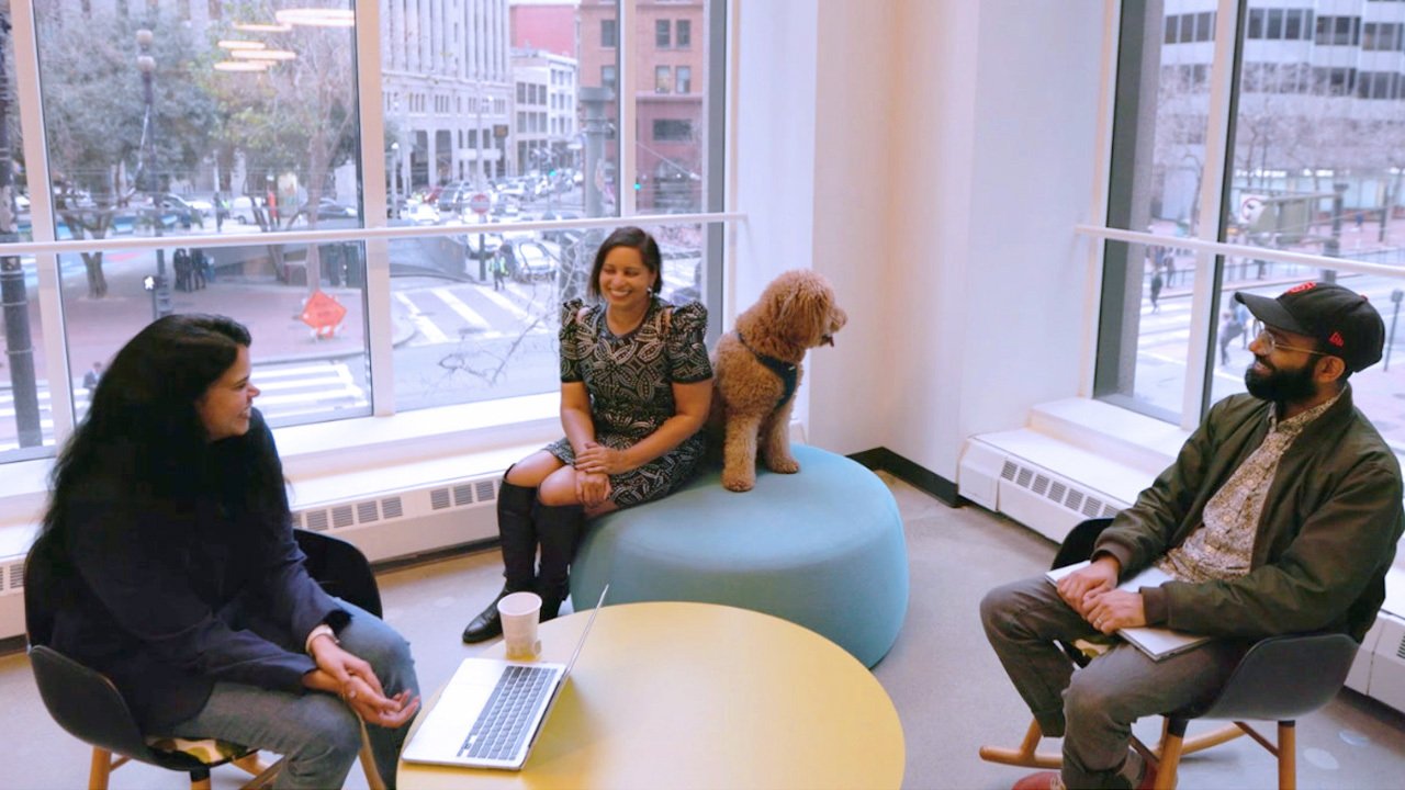 Three AWS AI/ML employees chat about their roles in Amazon's San Francisco office. They're sitting in front of office windows downtown, and one employee has a medium-sized dog sitting next to her.