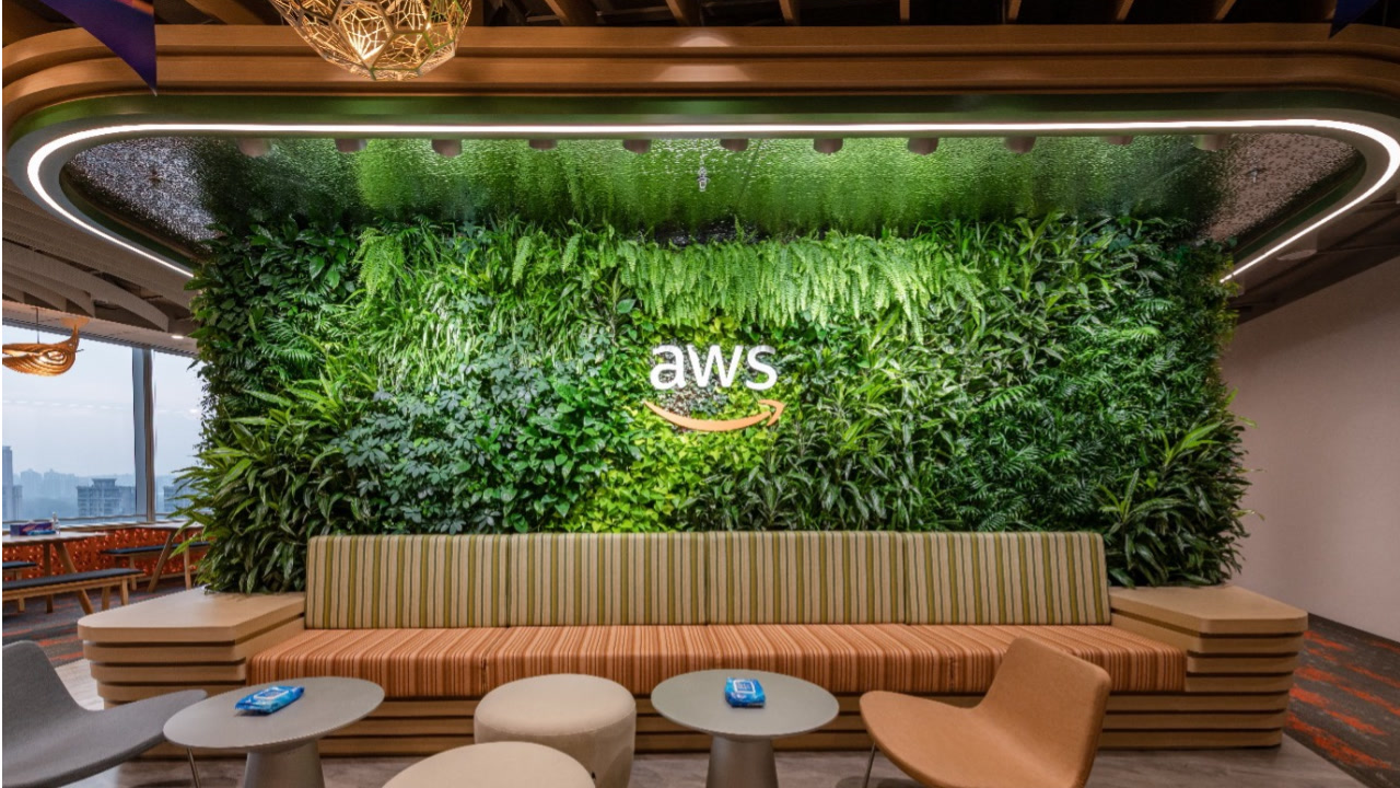 An image of the AWS logo in an Amazon office. It appears as a sign on a wall of live green plants, with a seating area for employees in front and beside it.