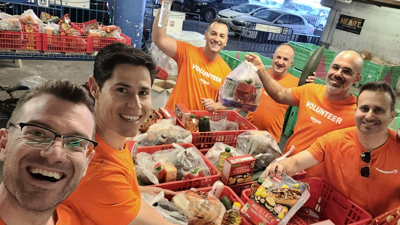 Amazon volunteers, wearing casual clothing and branded orange Amazon volunteer T-shirts, are working together to pack the boxes for distribution to approximately 500 families on behalf of the Petachon Lev association's poverty assistance programs.