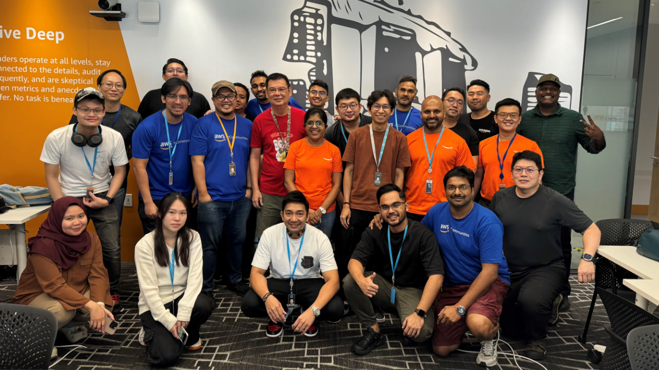 &quot;A group of around 20 Amazon employees, a diverse group of men and women, standing together and smiling at the camera after volunteering for a laptop refurbishment project. They are wearing casual clothing and are in an office setting.