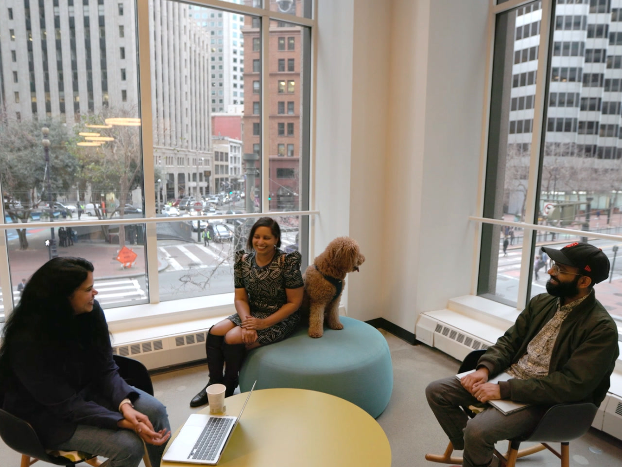 Three AWS AI/ML employees chat about their roles in Amazon's San Francisco office. They're sitting in front of office windows downtown, and one employee has a medium-sized dog sitting next to her.