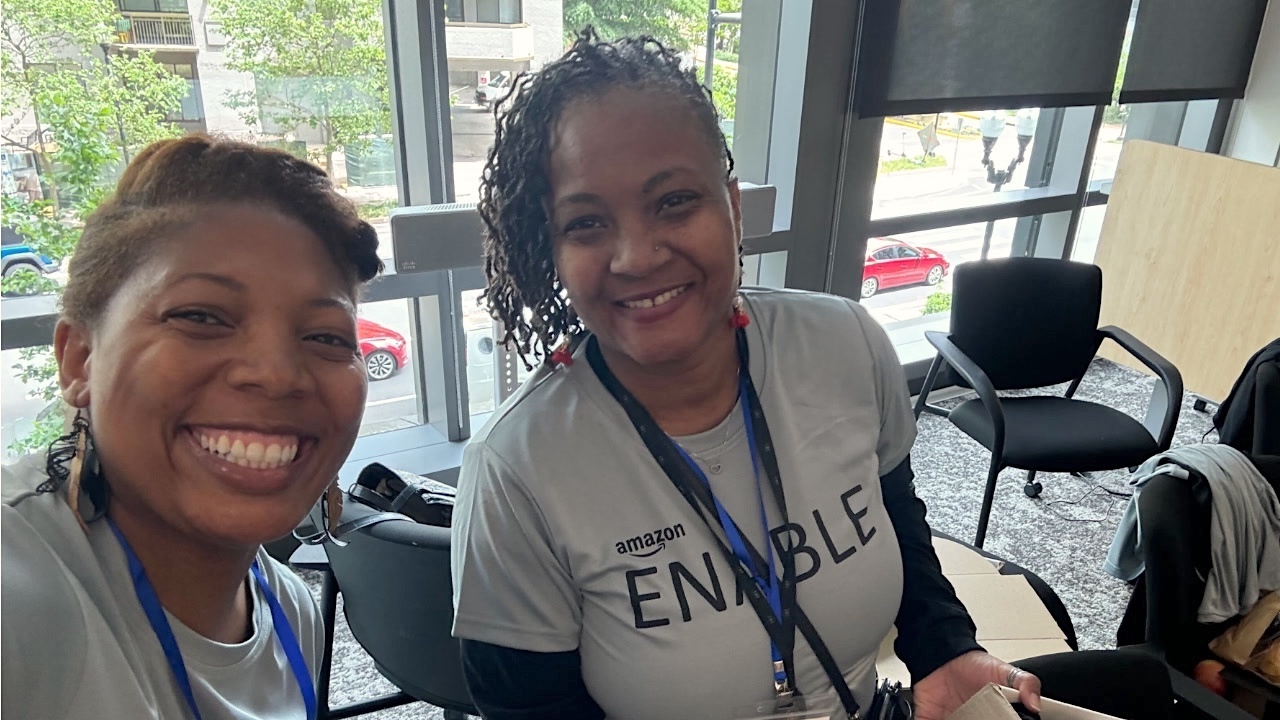 he image shows two smiling Black women Amazon executives standing together. They are wearing casual clothing and are posing for a photo at the ENABLE Summit, where they volunteered to build kits for the Central Union Mission, a nonprofit organization that supports individuals experiencing homelessness and poverty.