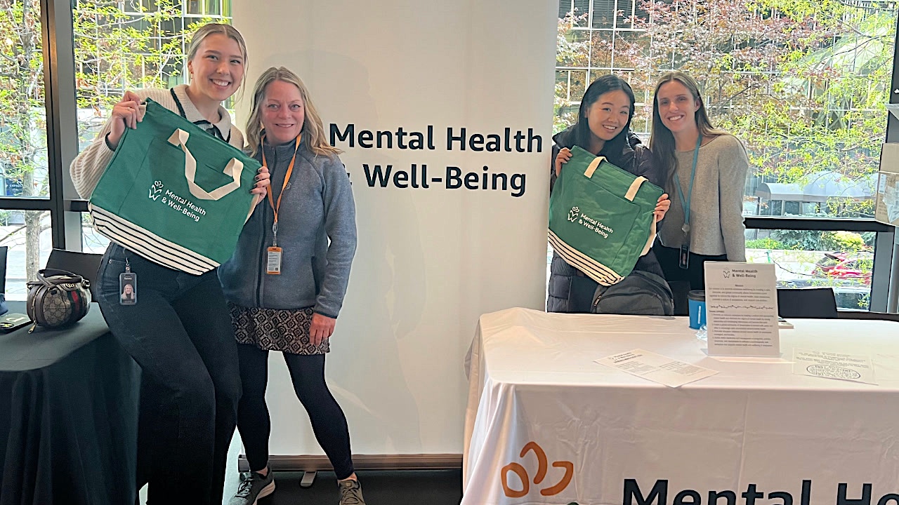 Four women Amazon employees post in front of a sign that says &quot;Mental Health and Well-Being. They are working at a booth at a sales event to promote the affinity group's work at Amazon. They are also holding up green bags with the affinity group's name and logo printed on them. 