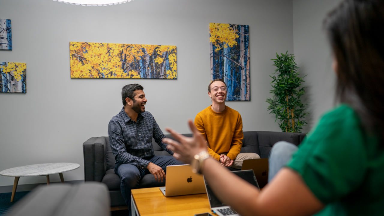 Three people sit around a table together with their laptops open, smiling and conversing with each other in an office setting. 