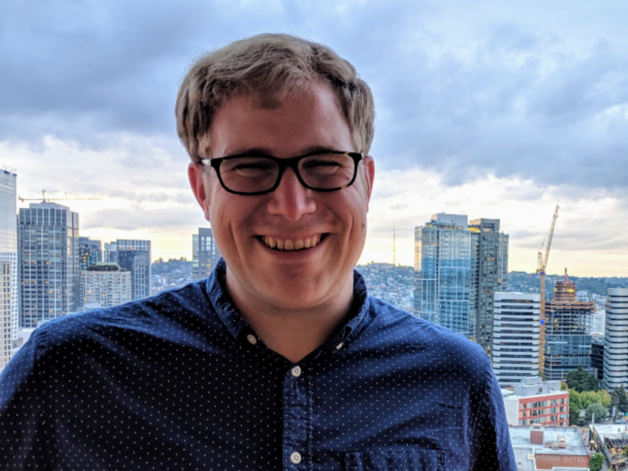 Steve Ash smiles for a portrait photograph outside with a city skyline in the background.