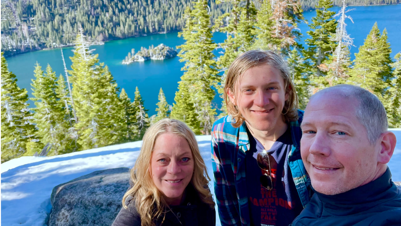 Julie Liveris, co-director of partnerships for the Mental Health and Well-being affinity group at Amazon, poses for a photo outdoors at Lake Tahoe with her husband and son. There are large fir trees behind them and a bright blue lake.