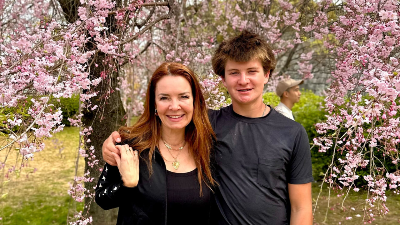 Anasuya Strasner poses outdoors in front of a blooming tree full of pink flowers. She is with her teenage son and they are smiling with their arms around each other.