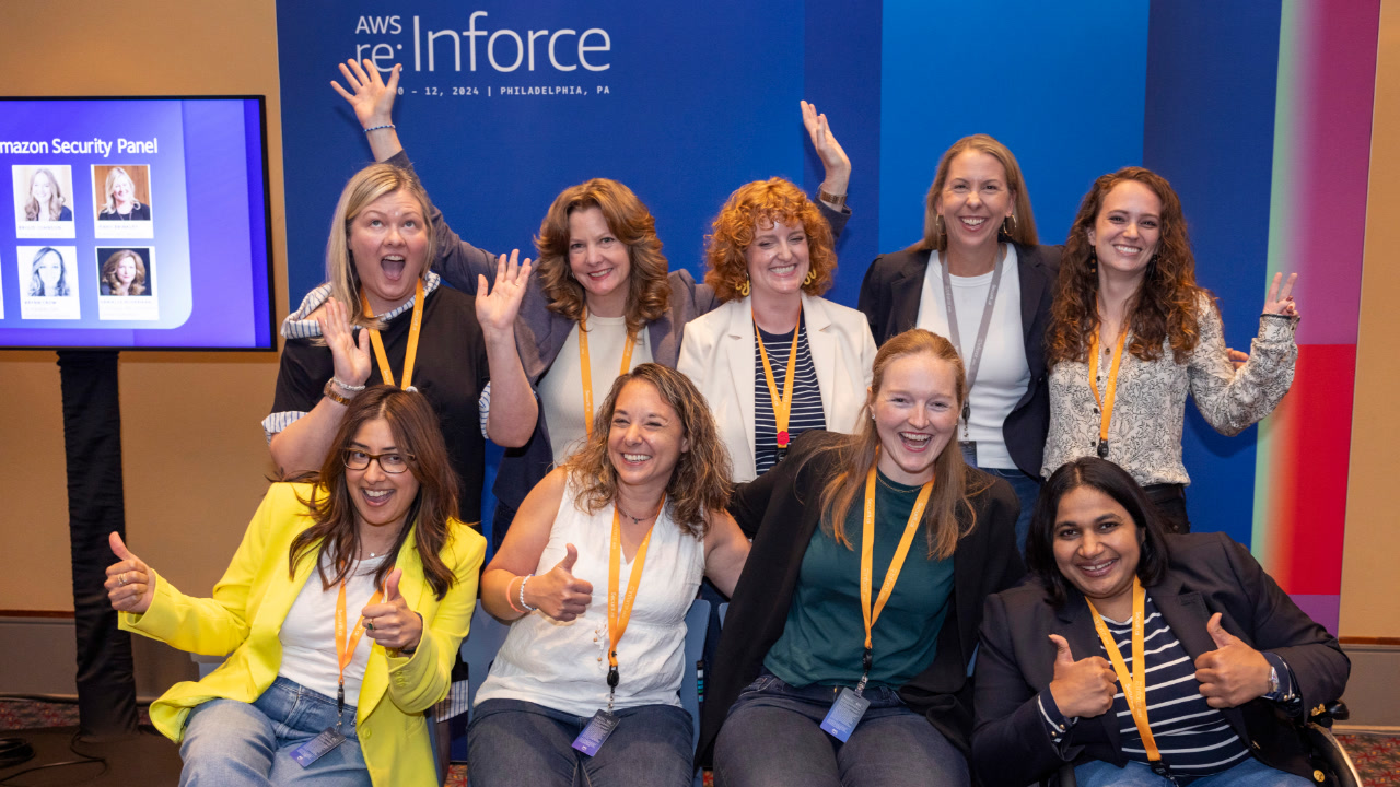 Nine women in security roles at Amazon and AWS smile for a group photo after participating in a panel discussion at AWS re:Inforce. On the wall behind them is an AWS re:Inforce logo. 