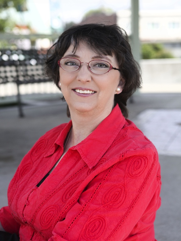 Charli Riggle, a principal accessibility program manager for AWS&rsquo;s software builder experience team and interim co-chair of Amazon&rsquo;s People with Disabilities affinity group. She is wearing a red blouse, round glasses, and smiling while posing from slightly to the side. The image is a portrait style image of Charli, who presents as a white woman, and was taken outdoors.