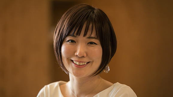 Shoko Utsnomiya, an AI/ML Specialist Solutions Architect with AWS, smiles for the camera against a brown background.