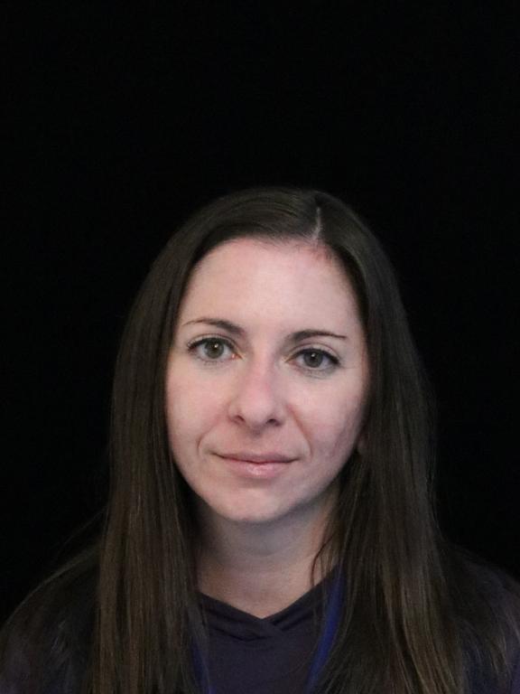 Justine Pascalides, head of accessibility at AWS, in her work photo. The image is a portrait-style headshot, showing Justine with a slight smile. She presents as a white woman with below-shoulder length brown hair. 