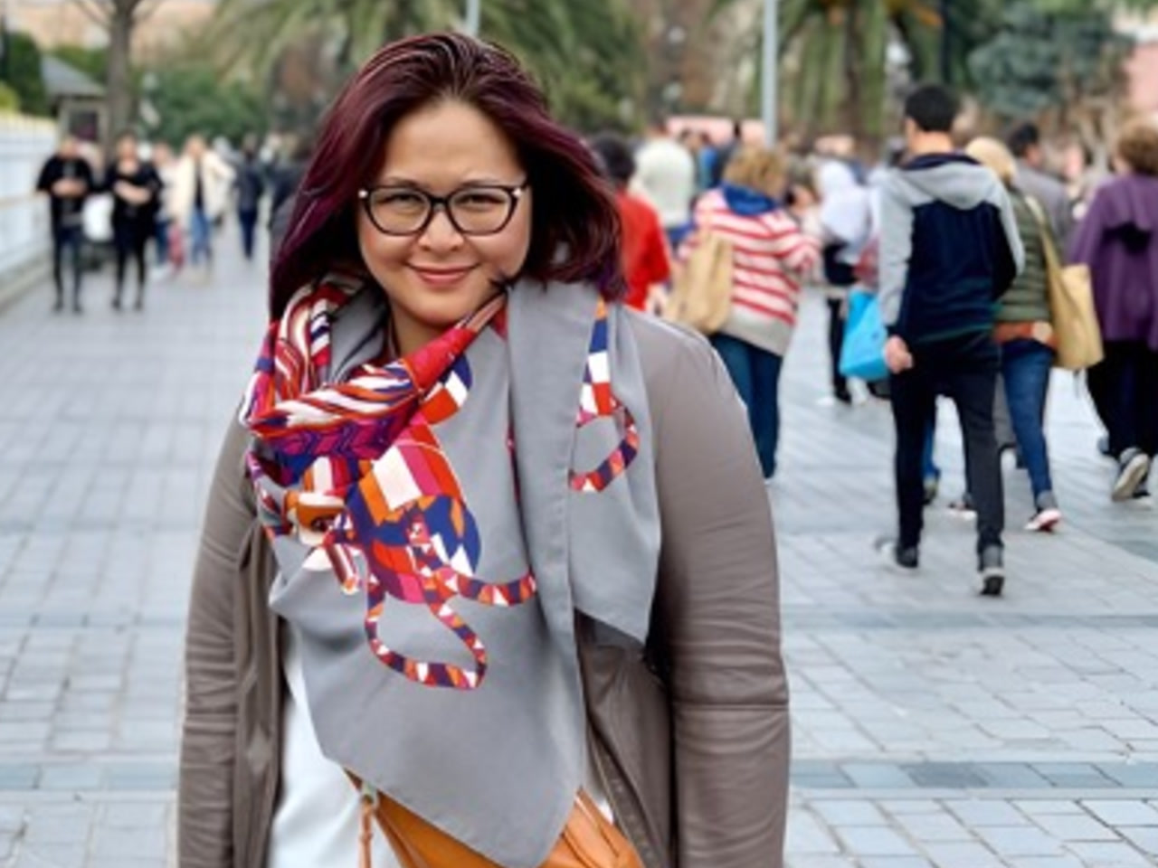 An image of AWS employee Amanda Kiemas, standing outdoors while many people walk in both directions behind her. She is smiling at the camera, wearing a bright colored scarf and black-frame glasses. 