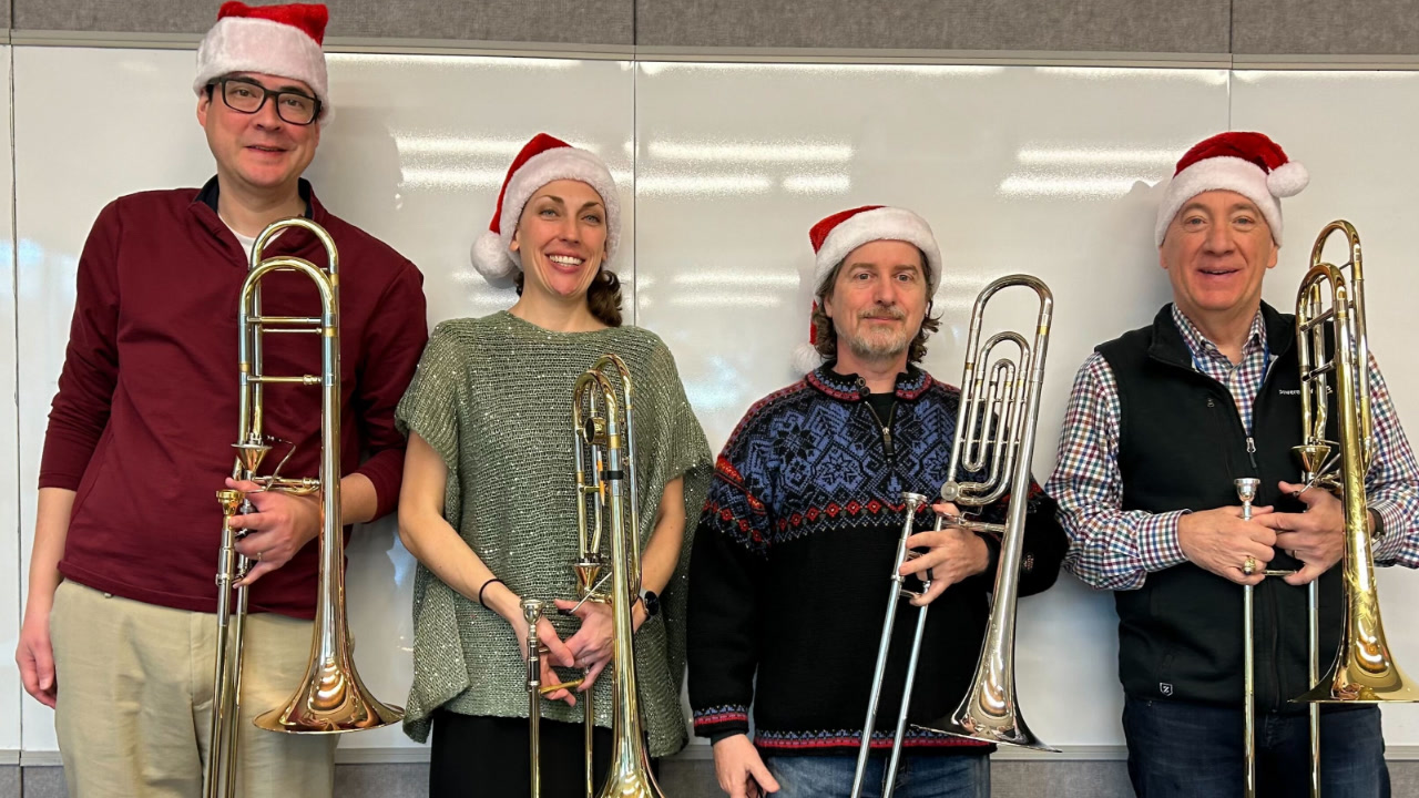 Four members of the AWS Trombone Quartet, standing in an AWS office against a whiteboard wall while holding their trombones. They are wearing Santa Claus hats after performing for the holidays. 