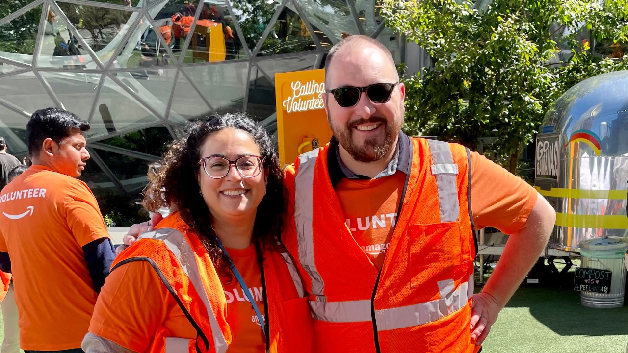 Two smiling Amazon employees, a man and woman wearing orange Amazon vests and volunteer T-shirts, pose together in front of the iconic Amazon Spheres glass domes in downtown Seattle. They volunteered together to assemble hygiene kits for people who have been displaced.