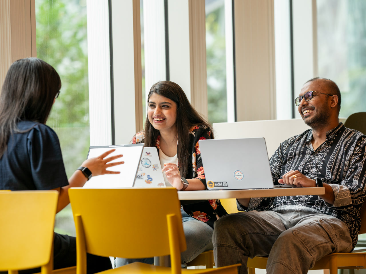 Three people sit around a table together with their laptops open, smiling and conversing with each other in an office setting. 