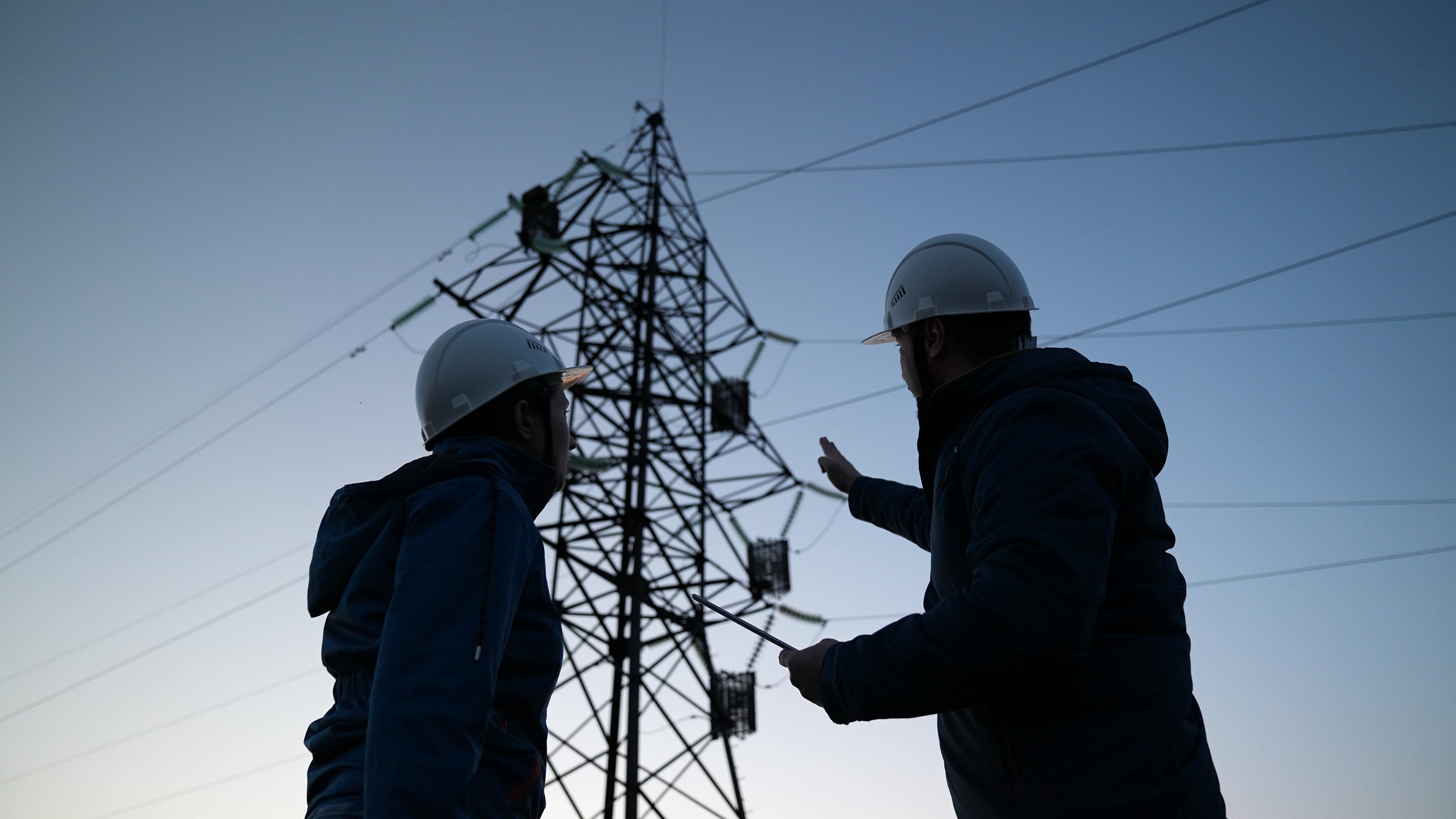 Power engineers are discussing the work plan. Two engineer standing on field with electricity towers at sunset. Silhouette of engineers looks at construction of high-voltage power. Engineering team