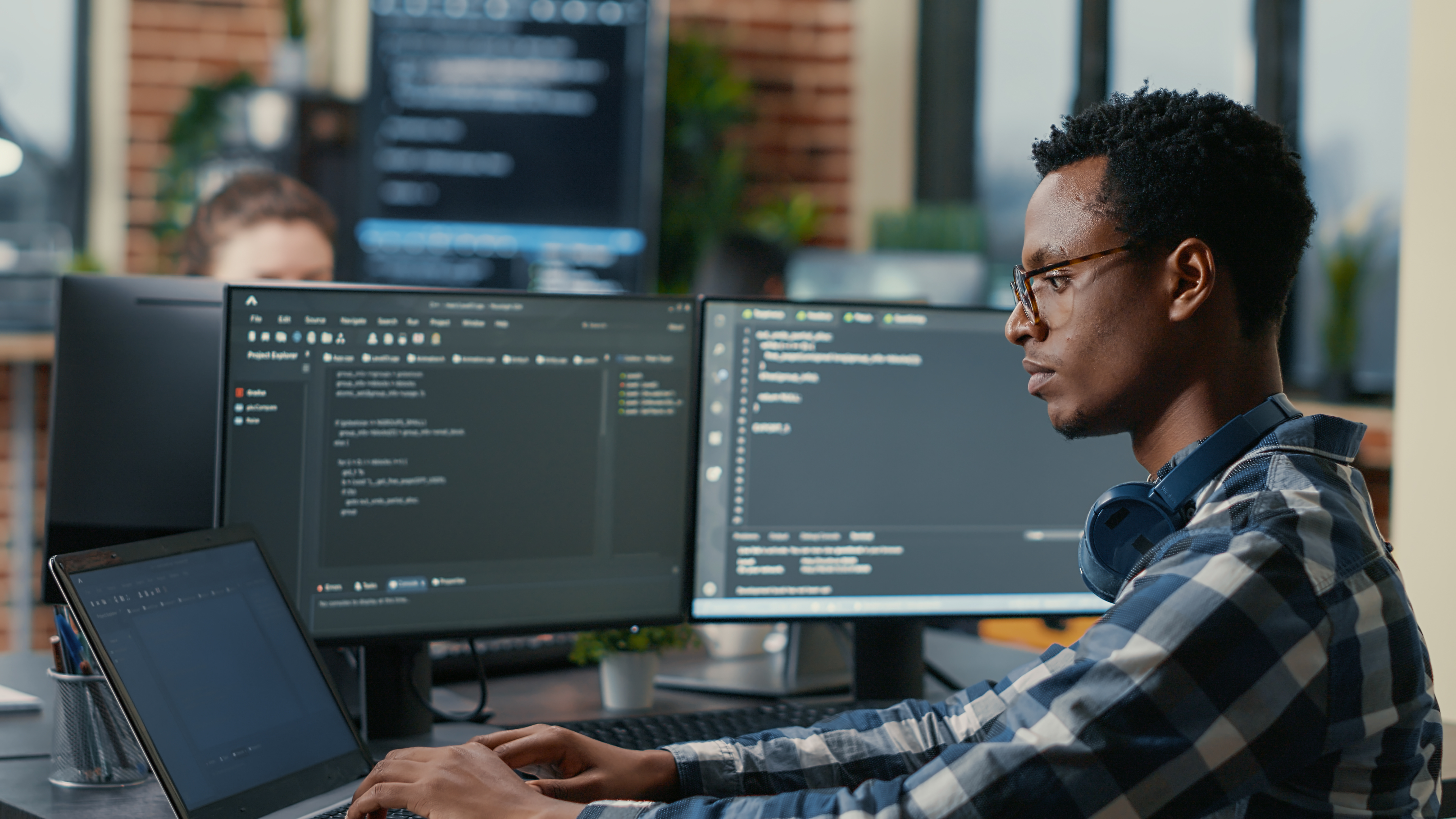 Sofware developer thinking while touching beard while typing on laptop sitting at desk with multiple screens