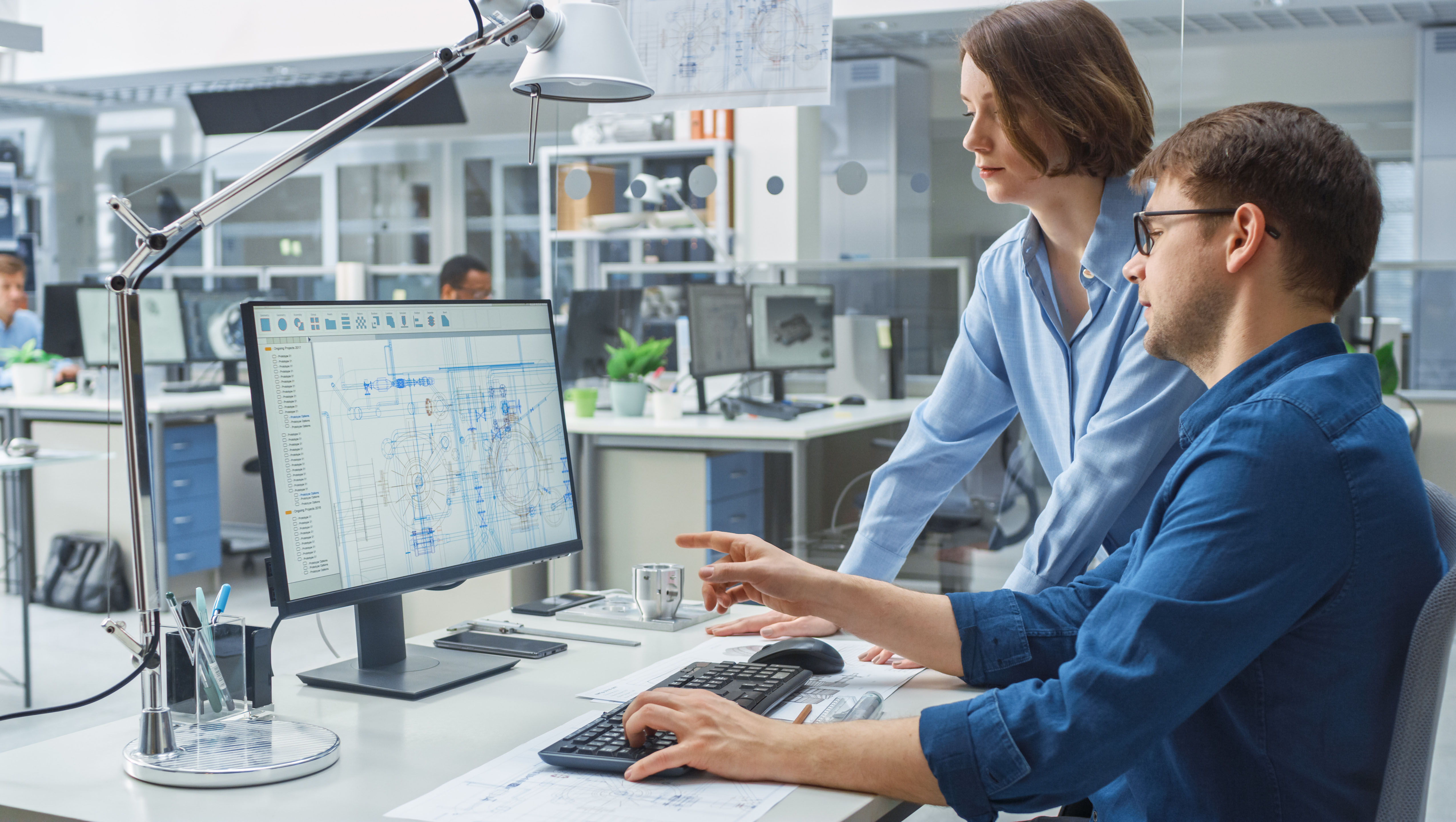 Engineer Talking With Project Manager and Working on Desktop Computer Using CAD Software with Technical Drawings on the Screen. In the Background Engineering Facility Specialising on Industrial Design