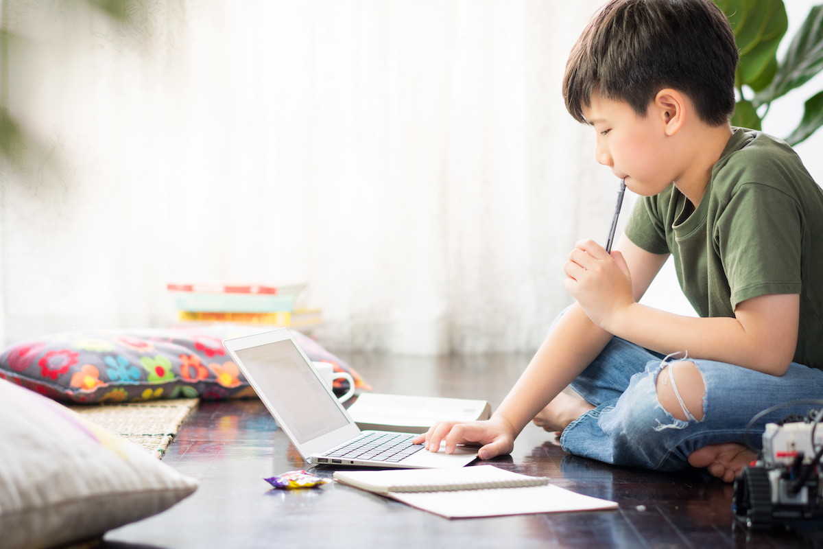 Smart looking Asian preteen boy sit crossed legs, holding pen against his lips, thinking and looking at computer laptop at home due to Covid-19 pandemic and social distancing measures