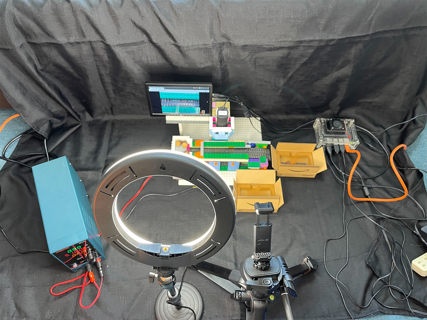 A tabletop machine learning experimental setup featuring a LEGO model, a ring light, camera tripod, electronic equipment, and a computer monitor, all arranged on a black cloth background.