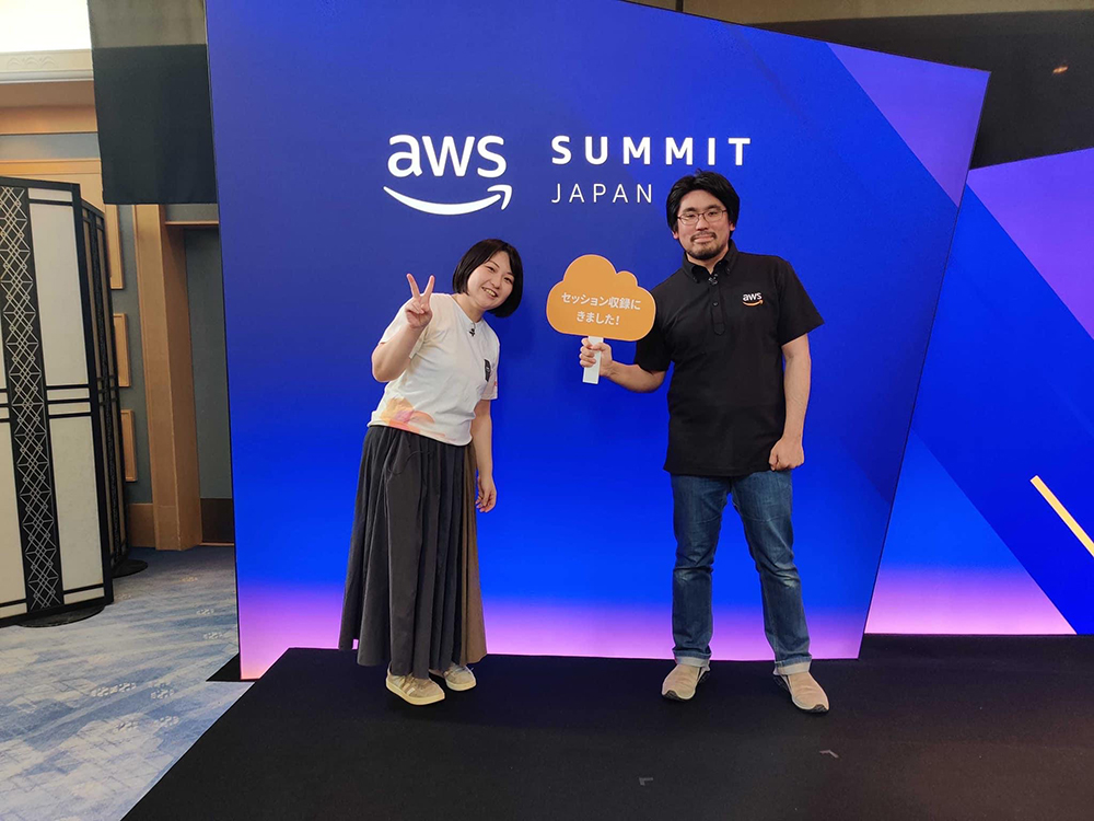 Two attendees pose for a photo at the AWS Summit Japan 2024 in front of an event backdrop. One attendee is holding a sign in Japanese, and the backdrop displays the AWS Summit Japan logo.