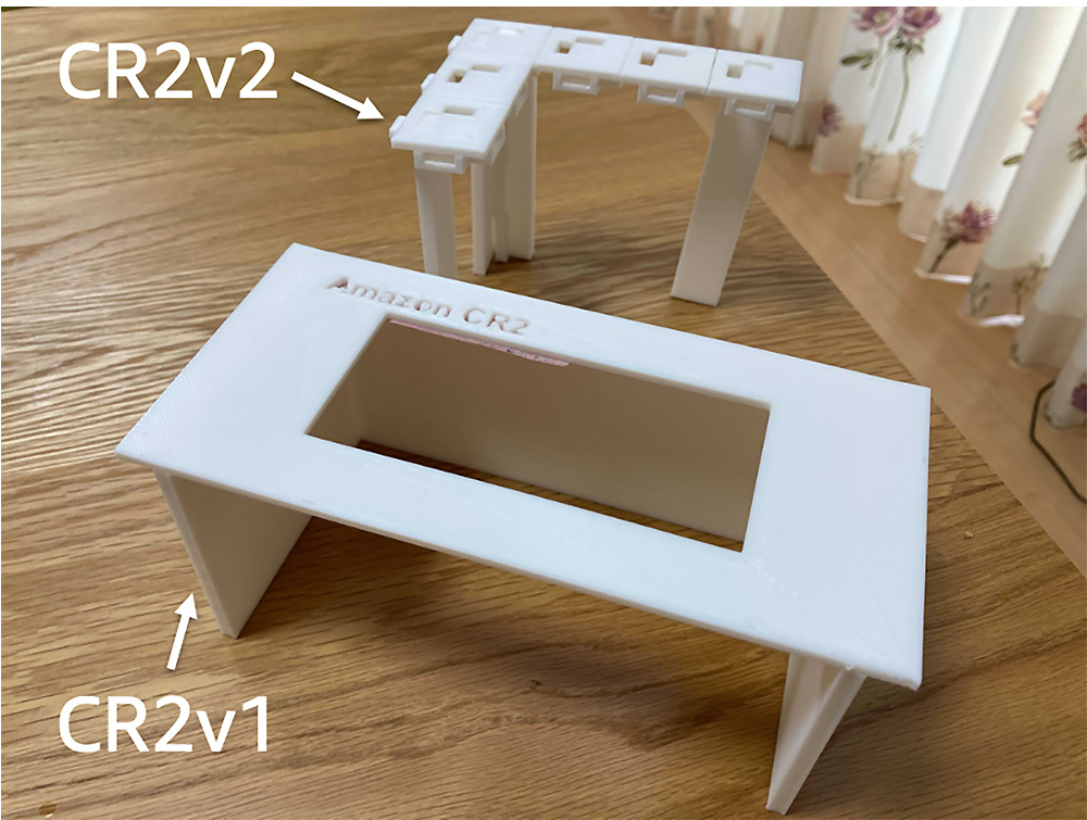 A comparison photo showing two 3D printed mounts labeled CR2v1 and CR2v2 for Amazon CR2, placed on a wooden surface.