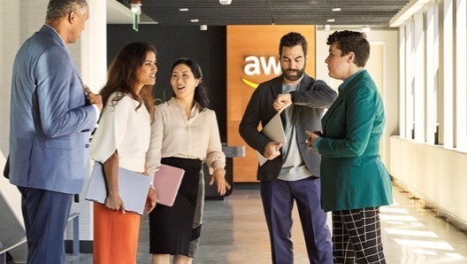 A diverse group of professionals having a discussion in an AWS office hallway, with the AWS logo visible in the background.