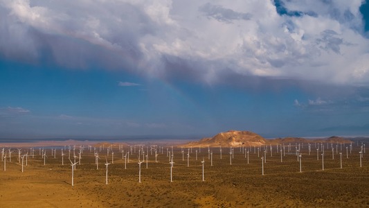 A wide view of a wind farm located in a desert landscape, with numerous wind turbines under a dramatic sky featuring a rainbow.
