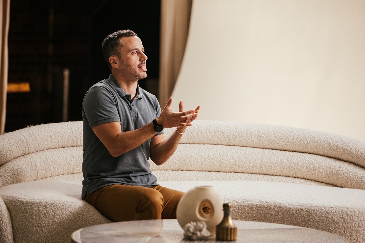A person seated on a textured white couch gesturing with their hands, with decorative objects on a table in the foreground.