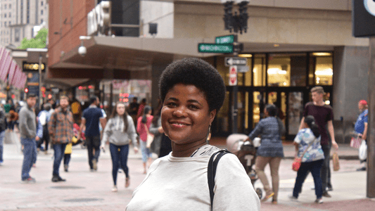 A woman smiling outdoors in an urban setting with people walking in the background. The photo is a headshot taken on a city street.