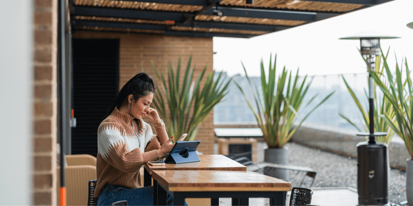 A woman sits at an outdoor table using a smartphone and a tablet with earbuds in, surrounded by plants and modern decor.