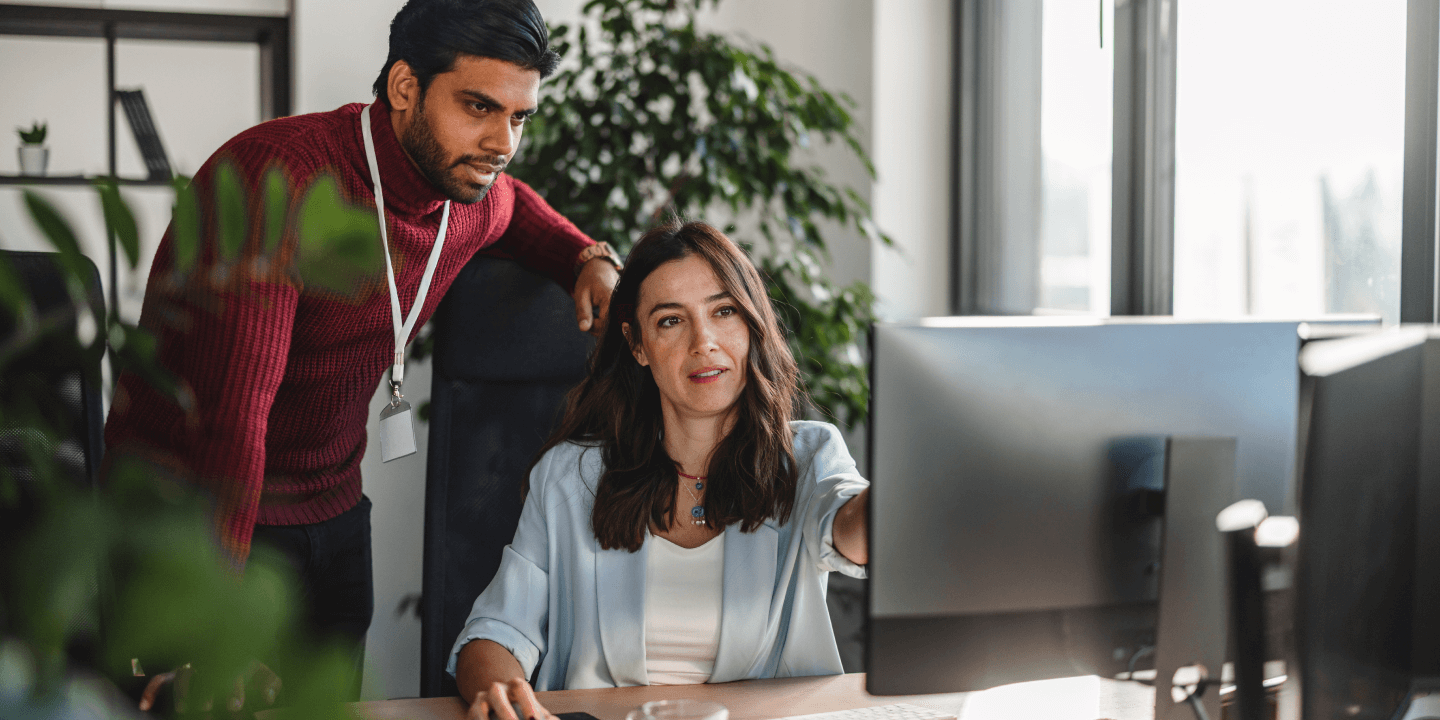 A woman seated at a desk points to a computer monitor while explaining something to a male colleague standing beside her in a modern office workspace.