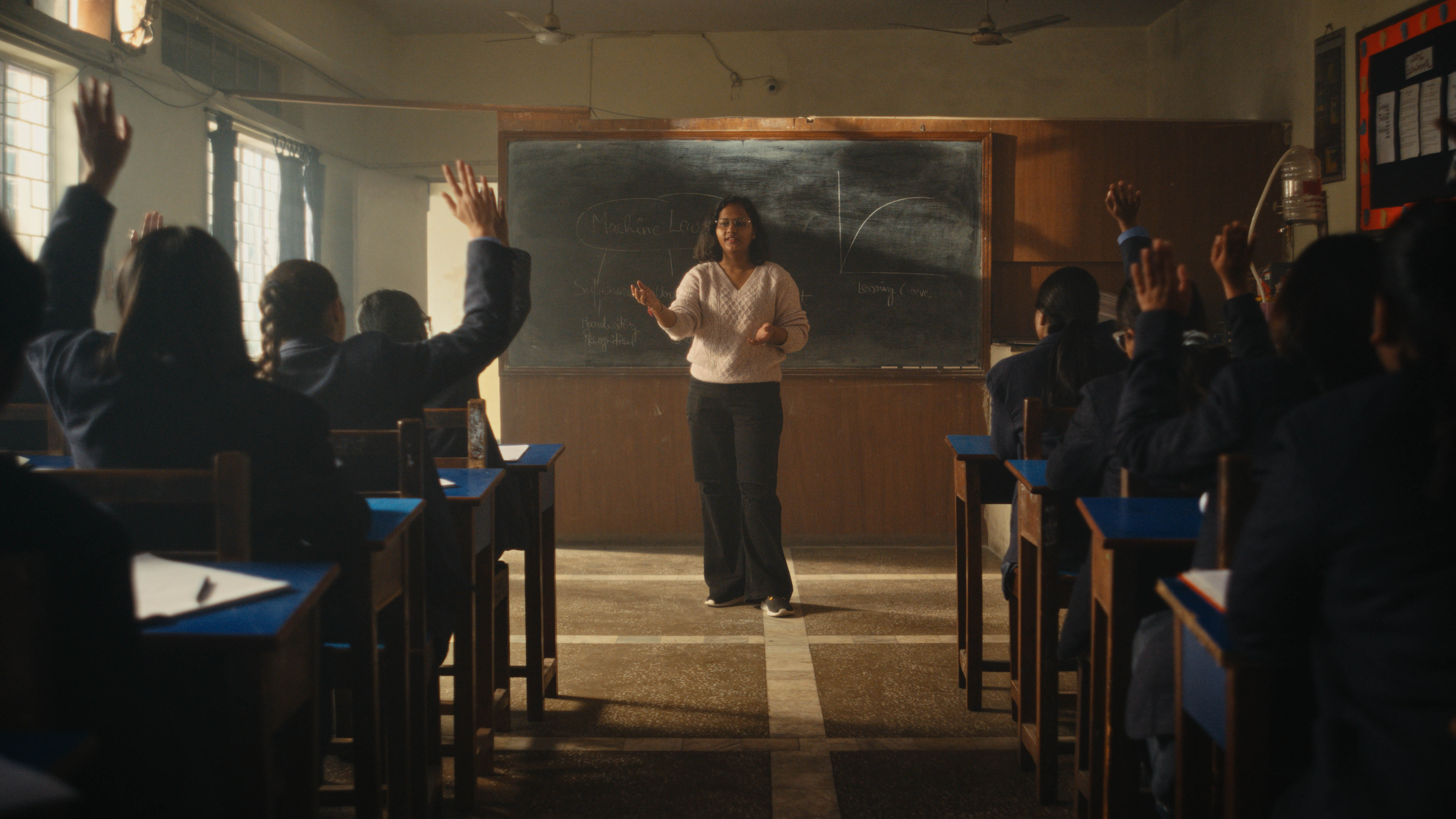 A teacher instructs a group of students in a classroom, with several students raising their hands to participate. The classroom has a blackboard and wooden desks, and the setting conveys active engagement in learning.