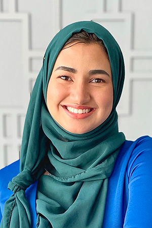 A professional headshot of a woman smiling, wearing a green hijab and blue blazer, with a geometric background.