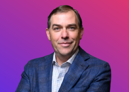 A professional portrait of a man in a checked shirt and blue blazer, set against a gradient purple and pink background, used for the Global Partner Summit.