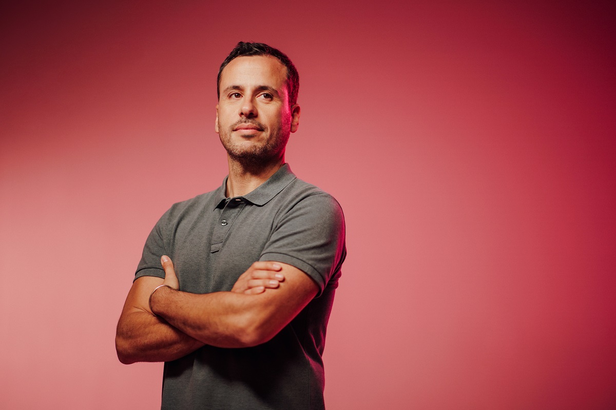A person in a gray polo shirt standing with arms crossed against a pink gradient background.
