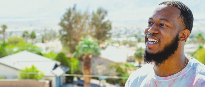 A man smiling outdoors with houses and trees in the background on a sunny day.