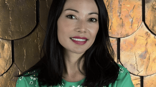 A professional headshot of a woman with long dark hair wearing a green dress, posed in front of a gold textured background.