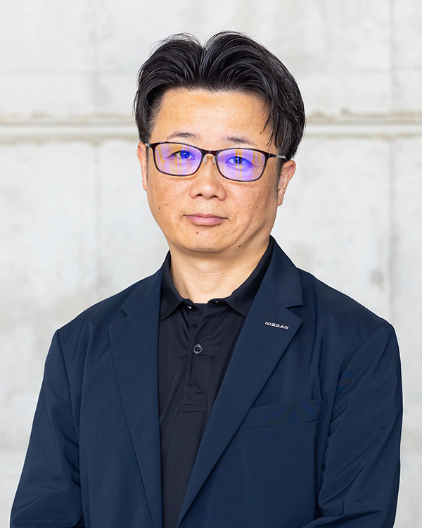 A person wearing a dark blue blazer with a Nissan logo and a black collared shirt, standing against a light concrete wall.