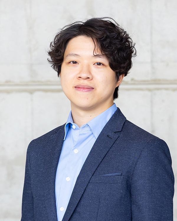 A person wearing a blue dress shirt and a dark blazer, standing in front of a light gray concrete wall.