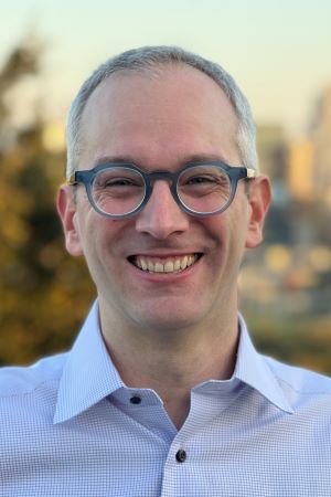 A portrait photo of a smiling person wearing glasses and a collared shirt, outdoors with a blurred background.