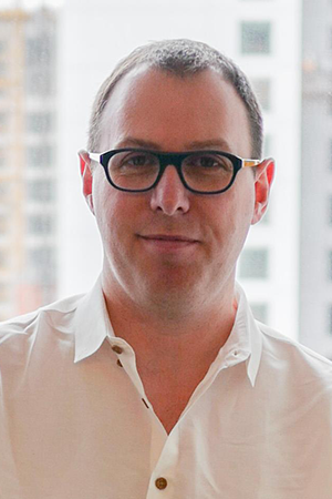 Portrait photo of an author wearing glasses and a white shirt, standing indoors, with blurred buildings in the background.