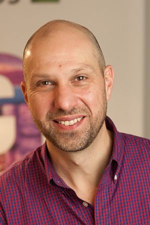 Portrait photo of Mark Mansour, author, smiling and wearing a red and blue checked shirt.