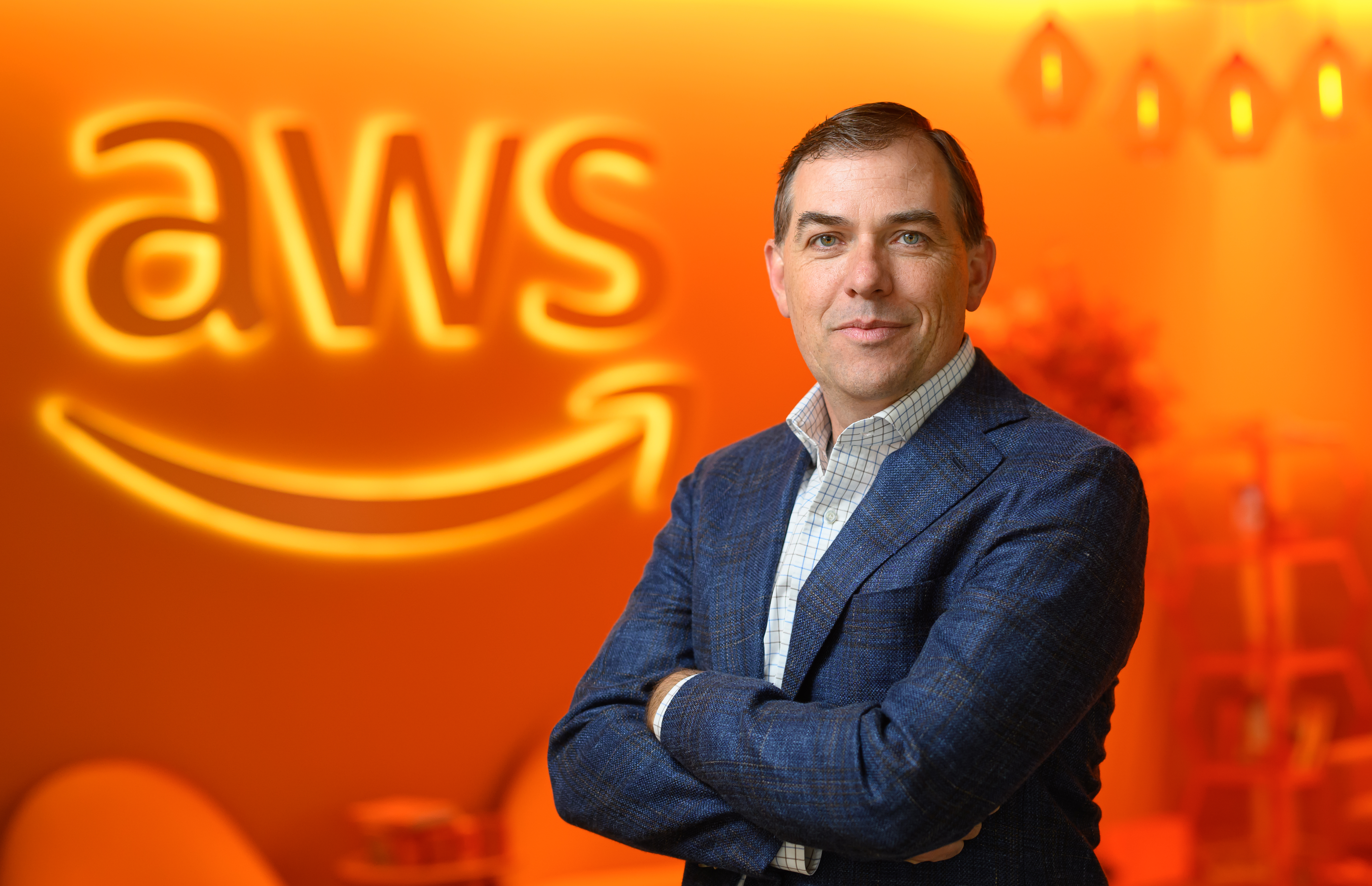 A person in a blue blazer standing in front of an orange background with the illuminated "AWS" logo.