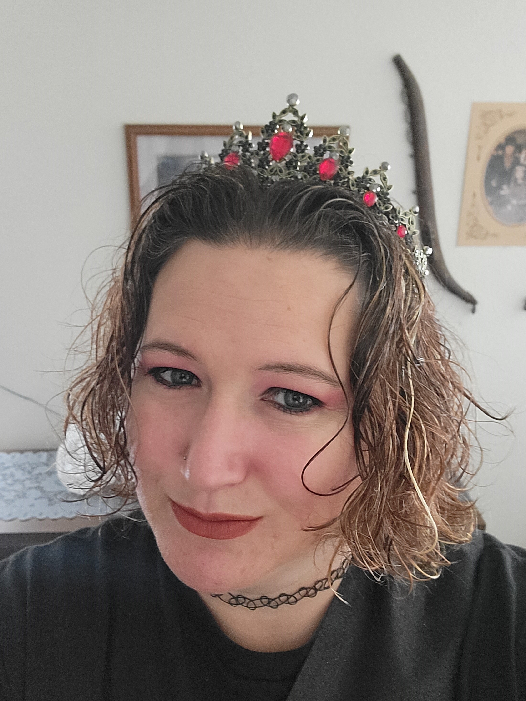 A woman with short, wavy hair is taking a selfie while wearing an ornate crown decorated with red gems. She is indoors, and the background shows framed pictures and household decor on the wall.
